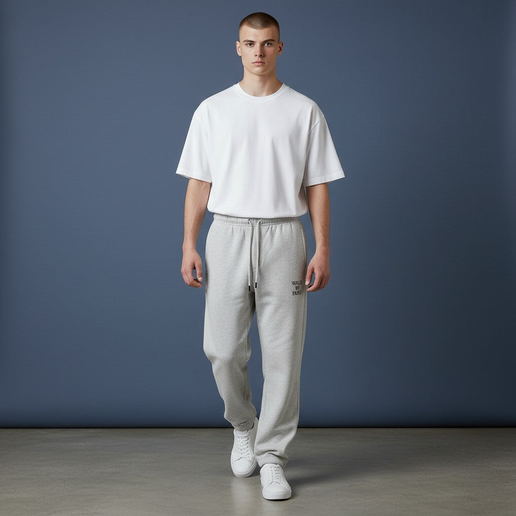 Omina Studio light grey cotton sweatpants with "Walk By Faith" text on the right thigh. Tapered fit, elastic waistband, drawstring, and cuffed ankles.
